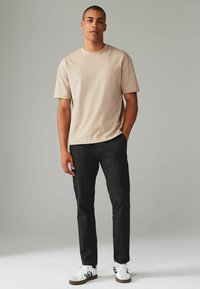 T-shirt beige à manches courtes avec un col rond, associé à un pantalon noir et des baskets blanches avec des accents noirs. Design simple et décontracté.