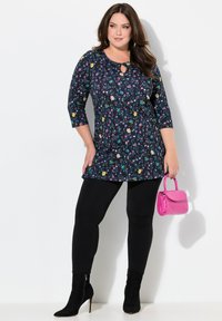 Blumen-Tunika mit Rundhalsausschnitt und Schlüsselloch-Detail, schwarze Leggings und einer kleinen pinken Handtasche. Merkmale einen Mix aus bunten Blumenmustern.
