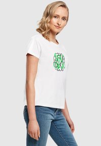 Hvid bomulds t-shirt med korte ærmer, prydet med et grønt og sort abstrakt design foran. Bæret med blå denim jeans.