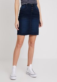 Tmavě modrá džínová sukně ke kolenům s přiléhavým designem, pět tlačítek vpředu a dvě přední kapsy. Spárováno s bílými vysokými teniskami.