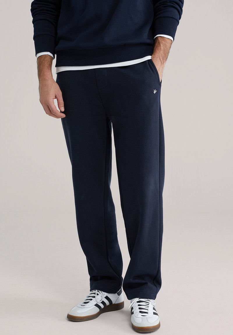 Tamno plave trenirke s elastičnim pojasom, bočnim džepovima i malim logotipom na kuku, uparene s bijelim tenisicama s crnim prugama.