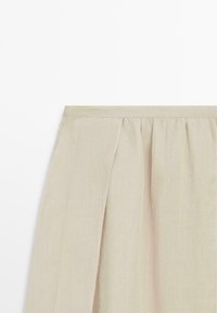 Beżowa lniana spódnica z marszczonym pasem i przednim rozcięciem, prezentowana na białym tle.