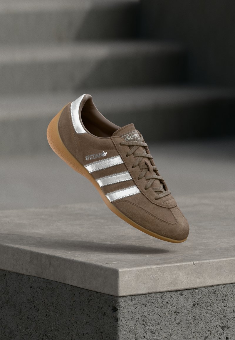 Καφέ σουέντ αθλητικό παπούτσι με λευκές ρίγες και ασημένιες λεπτομέρειες, με υφή στο πάνω μέρος, σχέδιο με κορδόνια και σόλα από καουτσούκ χρώματος καραμέλας.