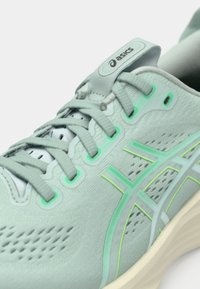 Sapatilha atlética cinza com parte superior texturizada, apresentando detalhes em verde claro e seções em malha. Os atacadores são planos, com colarinho e língua acolchoados.