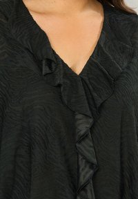 Blusa negra con un patrón texturizado, escote en V profundo y acentos con volantes en los bordes, fabricada con un tejido ligero.