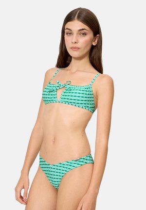 Mujer con cabello largo y castaño, vistiendo un bikini con patrón ondulado verde y blanco, con un detalle de lazo en la parte superior, de pie contra un fondo liso.