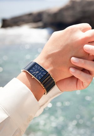 Rechteckige Uhr mit einem marineblauen Zifferblatt, silbernen Markierungen, goldfarbenen Akzenten und einem strukturierten schwarzen Armband. Am Handgelenk getragen vor einem natürlichen Hintergrund.