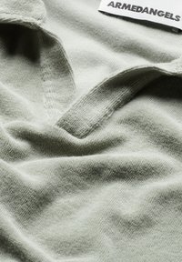 Helles grünes Baumwollgewebe mit einer weichen Textur und kleinem V-Ausschnitt. Verfügt über ein sichtbares Etikett mit dem Markennamen "ARMEDANGELS".