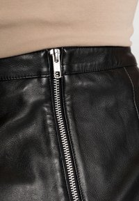 Jupe en cuir noire avec une fermeture éclair argentée à l'avant. Le matériau a une texture lisse avec des détails de couture subtils.