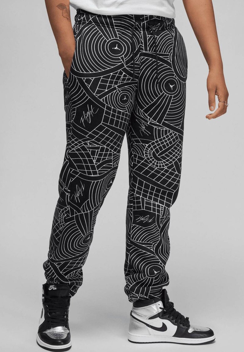 Zwarte joggers met een geometrisch witte lijnpatroon, voorzien van een elastische tailleband, zijzakken en een boord aan de enkels. Gecombineerd met zwarte en zilveren sneakers.