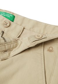 Béžové bavlněné kalhoty se zipem, dvěma knoflíky a zelenou visačkou s nápisem "United Colors of Benetton." Obsahují jemné prošívání a detaily loga.