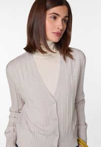 Hellbeige gerippter Cardigan mit V-Ausschnitt, Knopfverschluss und langen Ärmeln, über einem cremefarbenen Rollkragenpullover getragen. Weicher Strickstoff mit strukturierten Streifen.