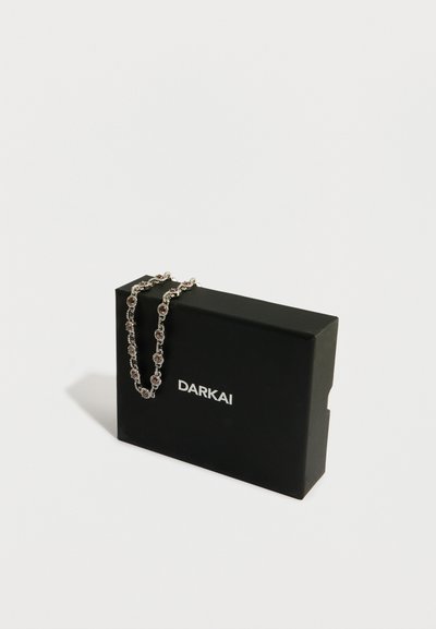 Srebrna verižica z okroglimi poudarjenimi kamni, razstavljena na črni škatli z oznako "DARKAI." Škatla ima mat zaključek in pravokotno obliko.