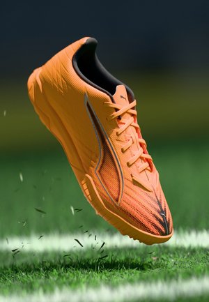 Zapatilla deportiva naranja con parte superior texturizada, detalles en negro y cordones contrastantes, suspendida sobre hierba verde, con escombros dispersos debajo.