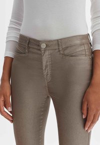 Braune Denim-Jeans mit schmaler Passform, ausgestattet mit Fronttaschen, einem Knopfverschluss und dezentem Glanz, kombiniert mit einem weißen Langarmoberteil.