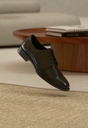 Chaussure habillée en cuir noir lévitant au-dessus d'un tapis beige tissé, avec une table basse ronde en bois et des livres empilés en arrière-plan flou.