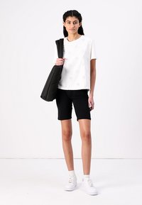 White short-sleeve top with floral cut-out pattern, paired with black bike shorts and white sneakers. A black shoulder bag is slung over one shoulder.