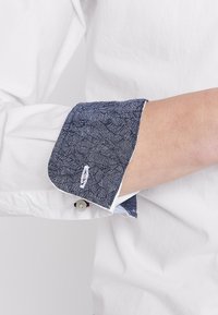 Manche de chemise blanche retroussée avec un revers intérieur contrasté bleu foncé à motif et une boutonnière visible sur l'avant-bras d'une personne.