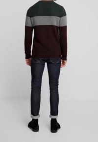 Gestrickter Pullover in Grün, Grau und Burgund-Streifen, kombiniert mit dunklen Denim-Jeans. Der Pullover verfügt über einen strukturierten Stoff und gerippte Bündchen.