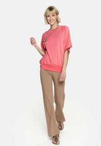 Koronkowa, oversize krótka bluzka w kolorze koralowym z szerokim dekoltem, połączona z beżowymi spodniami w stylu dzwonów oraz brązowymi sandałami na ramiączkach. Prosty detal w postaci srebrnego naszyjnika.