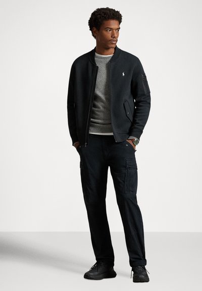 Čierna bombera bunda cez sivý sveter, v kombinácii s čiernymi cargo nohavicami a čiernymi teniskami. Obsahuje detaily so zipsom a vreckami.