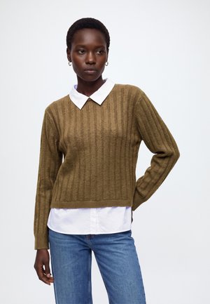 Femme aux cheveux courts portant des boucles d'oreilles créoles dorées, un pull côtelé vert olive sur une chemise blanche à col et un jean bleu, debout devant un fond blanc.