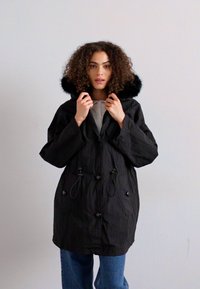 Black parka jacket with a faux fur-lined hood, drawstring waist, and two front pockets. Textured fabric with a relaxed fit, long sleeves.