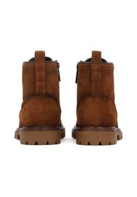 Stivali ankle in suede marrone con una texture liscia, dotati di suola in gomma spessa e di un'etichetta di trazione sul retro. Dettagli con zip sui lati.