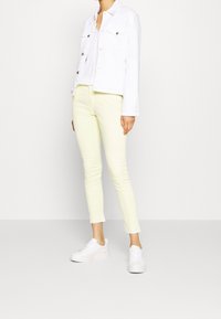 Giacca di jeans bianca, jeans skinny giallo chiaro e sneaker bianche. La giacca presenta due tasche frontali e bottoni metallici.