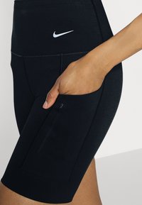Main dans la poche d’un short de sport noir Nike avec une poche latérale zippée et un logo Nike blanc sur la ceinture.