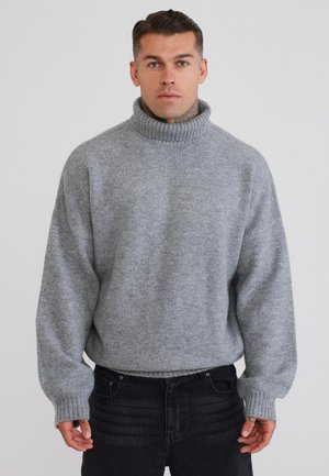 Mand iført en grå rullekrave og sorte jeans står mod en ensfarvet lys baggrund og vender fremad.