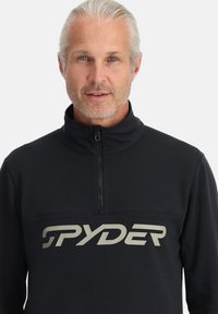 Pullover in pile nero con colletto alto e mezzo zip. Presenta un grande logo argento "SPYDER" sul petto. Texture morbida.