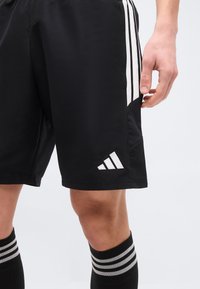 Άνδρας που φοράει μαύρα αθλητικά σορτς Adidas και μαύρες κάλτσες με λευκές ρίγες, στέκεται μπροστά από ένα απλό φωτεινό φόντο.