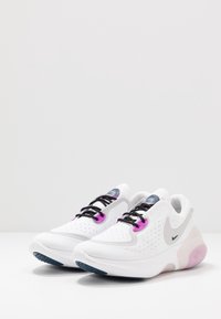Chaussures de sport blanches avec lacets noirs, accents violets et dessus texturés. Dotées d'une semelle coussinée avec des touches de rose et d'un logo de marque.
