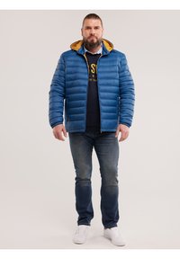  szakállas férfi kék pufi dzsekiben, sárga kapucnin bélés, sötét pulóver, kék farmer és fehér sportcipő, egyszínű háttér előtt állva.