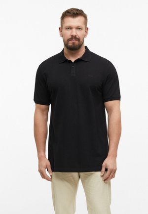 Homme barbu portant un polo noir à manches courtes et un pantalon beige, debout devant un fond blanc uni.