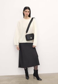 Suéter crema cómodo, falda midi de satén negra, botines negros con puntera brillante y un bolso de hombro estructurado negro con una correa decorativa.