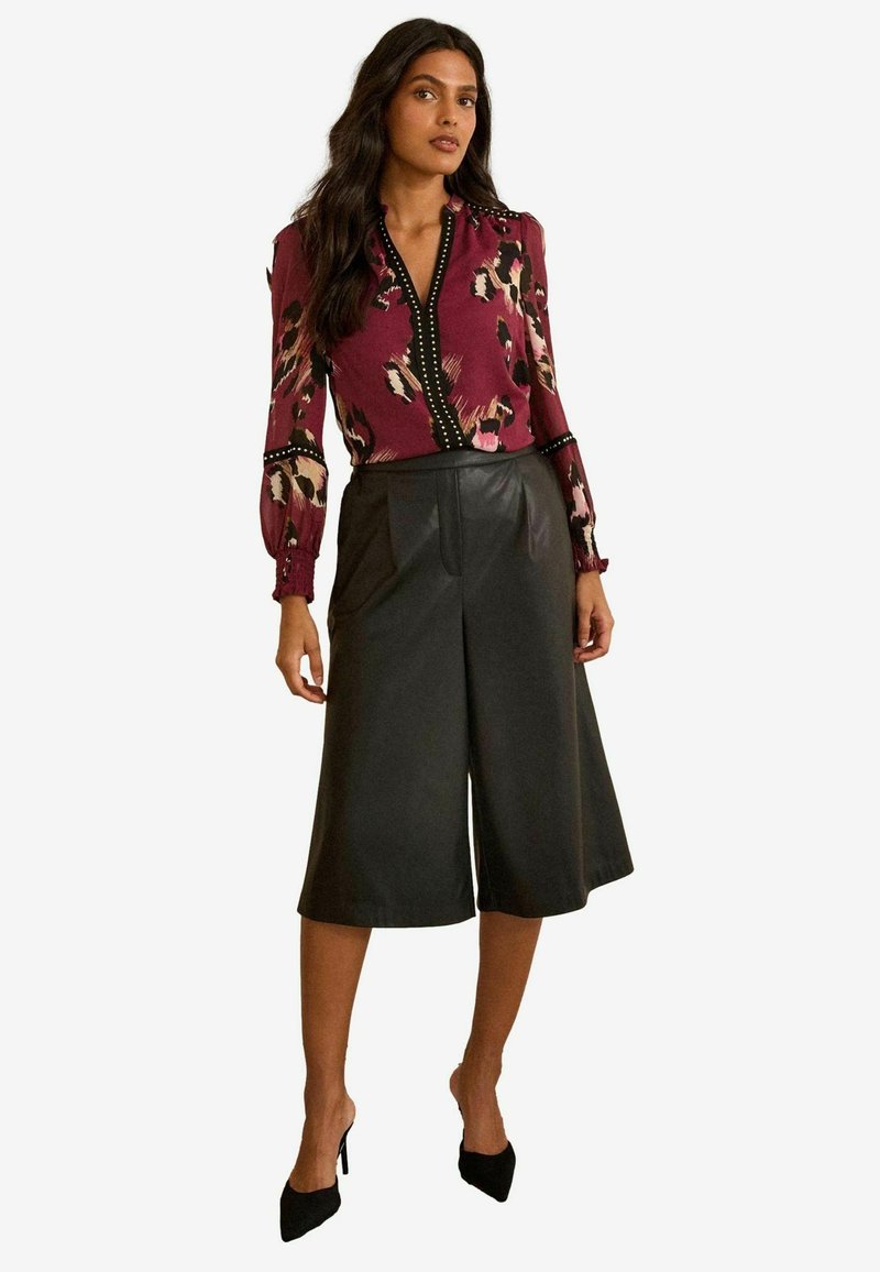 Langärmlige kastanienfarbene Bluse mit Tiermotiv und schwarzen Details, kombiniert mit hoch taillierten schwarzen Leder-Culottes, getragen mit schwarzen Absätzen.