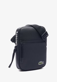 Bolso mensajero Lacoste negro con superficie texturizada, correa ajustable, cierre con cremallera y un pequeño logo bordado en la parte frontal.