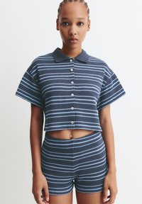 Une jeune femme avec des cheveux tressés portant un crop top à manches courtes boutonné aux rayures marine et bleu clair assorties, ainsi que des shorts taille haute.