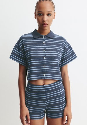 Junge Frau mit geflochtenen Haaren, die ein passendes kurzärmeliges, navy- und hellblau gestreiftes, knöpfbares Cropped-Shirt und hochgeschnittene Shorts trägt.