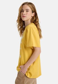 Geel katoenen t-shirt met korte mouwen, voorzien van een wit bedrukt ontwerp. Het shirt heeft een relaxte pasvorm en een gladde, zachte textuur.
