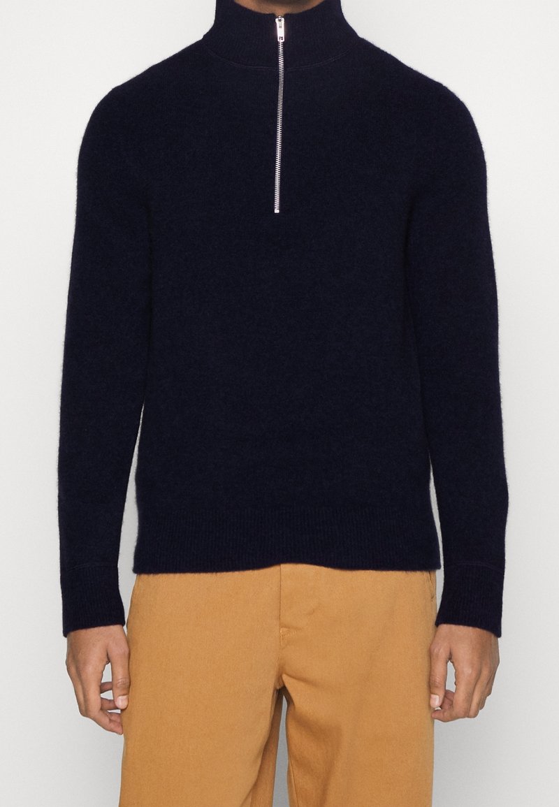 Homme portant un pull à manches longues bleu marine avec col à fermeture éclair partielle et pantalon couleur camel, debout devant un fond clair uni.