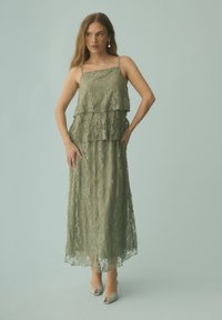 Femme en robe en dentelle à volants vert olive avec fines bretelles et talons ouverts beige, debout sur un fond uni gris clair.
