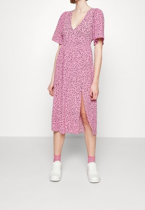 Kvinde iført en lyserød blomsterprint midi-kjole med korte ærmer, dyb V-hals, sideslids, lyserøde sokker og hvide sneakers, stående på en grå gulv.