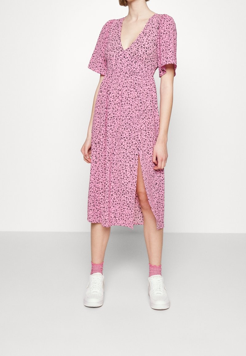 Femme portant une robe midi rose à fleurs avec manches courtes, décolleté en V profond, fente latérale, chaussettes roses et baskets blanches, debout sur un sol gris.
