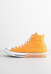 Vysoké tenisky v jasně oranžovém plátně, s bílými tkaničkami a gumovými podrážkami s černými pruhy. Hladká textura a klasický design.