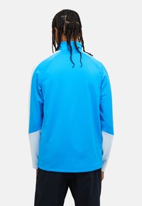 Giacca atletica blu a maniche lunghe con maniche raglan, colletto alto e dettagli bianchi, realizzata in materiale leggero e liscio.