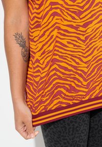 Camiseta de manga corta con un patrón de rayas de tigre en naranja y burdeos, que presenta un dobladillo acanalado con dobles rayas. Mano reposando en el costado.