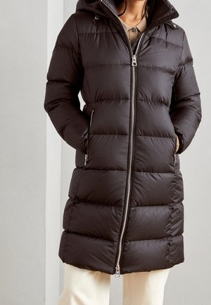 Person iført en lang sort dunjakke med hætte, lynet op, hænderne i sidelommerne, kombineret med lysfarvede bukser.